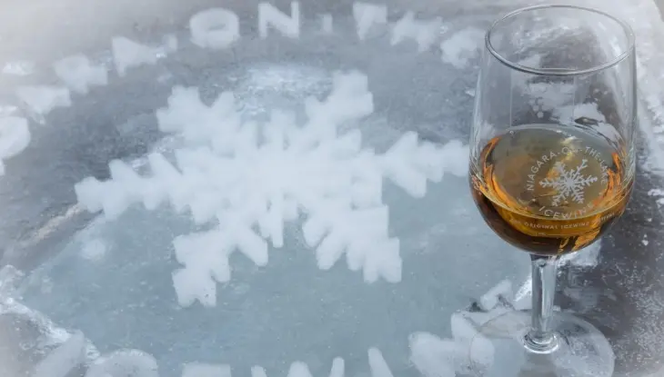 Ein mit bernsteinfarbener Flüssigkeit gefülltes Weinglas steht auf einer Eisfläche, in die eine Schneeflocke und die Worte „Niagara On The Icewine“ eingraviert sind.