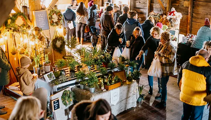 Visitors browsing plant displays and handmade goods at a market inside a rustic wooden barn.