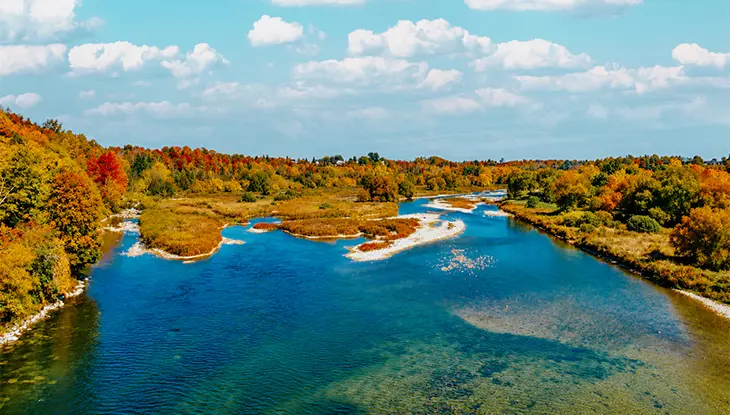 Vue aérienne d’une rivière bleue sinueuse traversant une forêt à l’automne, avec un feuillage vibrant aux teintes d’orange, de jaune et de rouge et de petits rapides entourant des îlots rocheux.