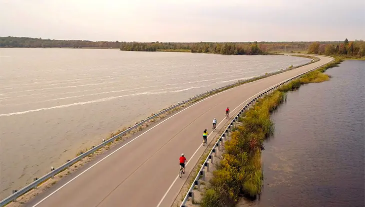 Aerial view of cyclists riding along a road between two bodies of water, with wetlands and shoreline stretching into the distance.