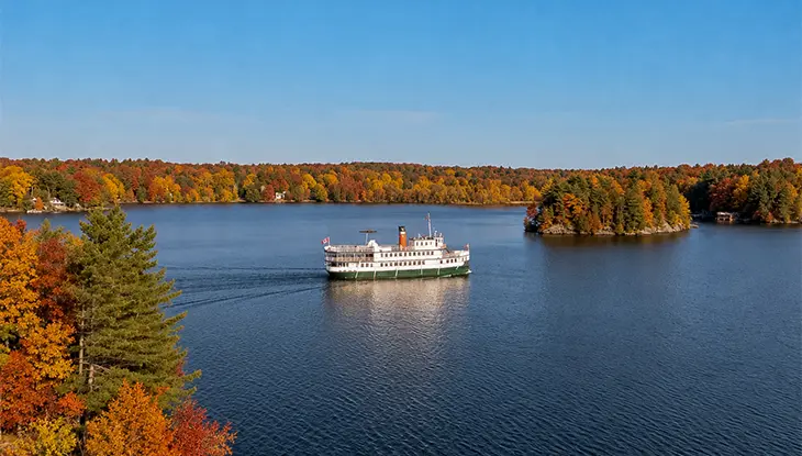 Ein Passagierboot gleitet über einen ruhigen See, umgeben von farbenprächtigem Herbstlaub und bewaldeten Inseln unter klarem blauen Himmel.