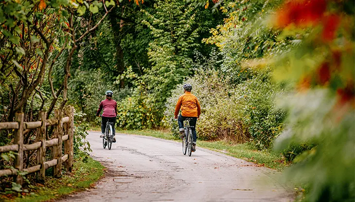 Two cyclists ride along a winding paved trail through a wooded area, bordered by dense greenery and a rustic wooden fence.