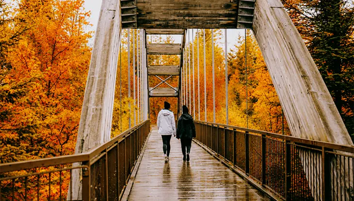 Two people walking across a wet bridge with orange fall trees surrounding them
