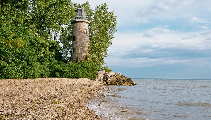 Phare en pierre entouré d’arbres, situé au bord d’un littoral rocheux.