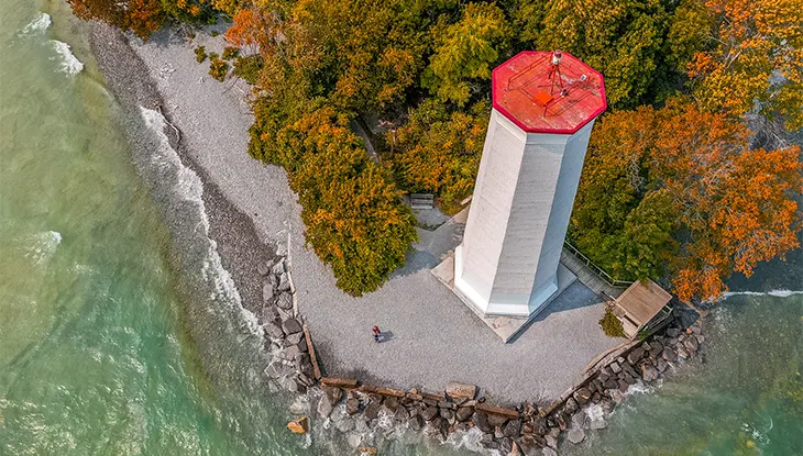 Vista aérea de un faro rojo y blanco situado junto a una costa rocosa con aguas cristalinas de color turquesa y follaje otoñal.