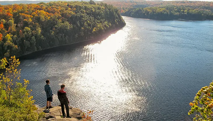 K33 Dos personas de pie en un mirador rocoso con vistas a un lago iluminado por el sol y rodeado de bosques otoñales.