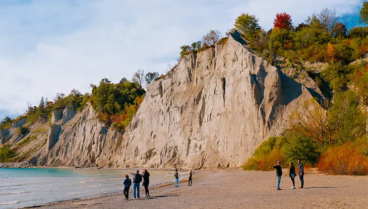 People walk along a sandy beach below towering white cliffs beside a lake.