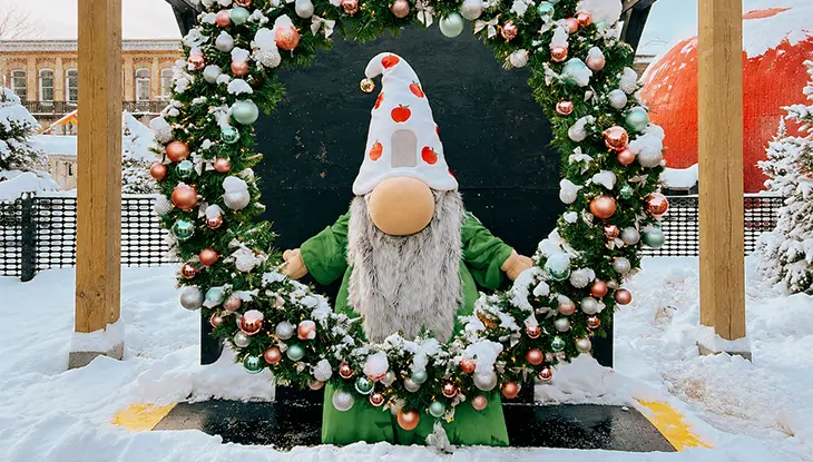 A gnome in a green outfit and apple-patterned hat stands inside a snow-covered Christmas wreath decorated with colourful ornaments and lights.