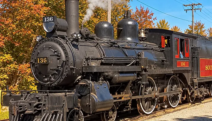 Eine historische Dampflokomotive fährt auf einer Eisenbahnstrecke, umgeben von Bäumen mit leuchtendem Herbstlaub.