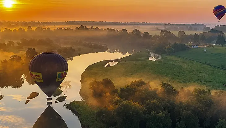 Heißluftballons schweben bei Sonnenaufgang über einem gewundenen Fluss und einer bewaldeten Landschaft, warmes goldenes Licht spiegelt sich im Wasser.