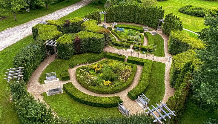Un jardín formal visto desde arriba, con parterres geométricos, senderos sinuosos y setos bien cuidados rodeados de exuberante vegetación.