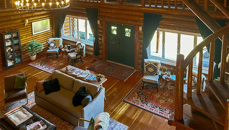 Interior rústico de cabaña con vigas de madera a la vista, chimenea de piedra, cómodos asientos y grandes ventanales con vistas al paisaje circundante.