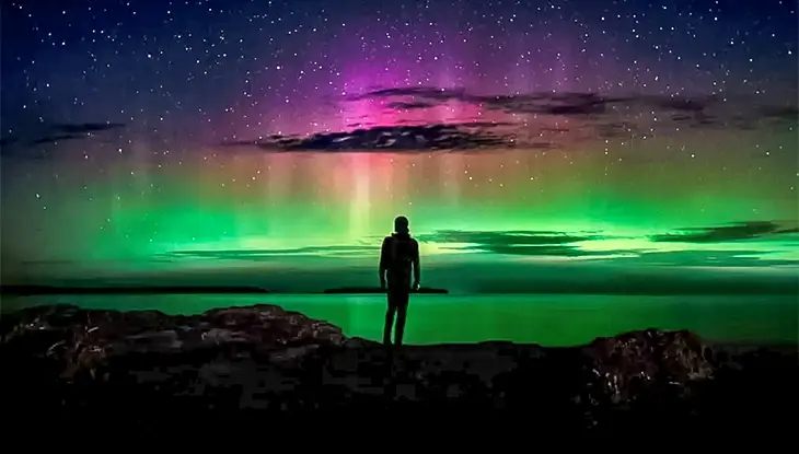 Une personne debout sur une rive rocheuse sous un ciel nocturne illuminé par des aurores boréales vertes et violettes.