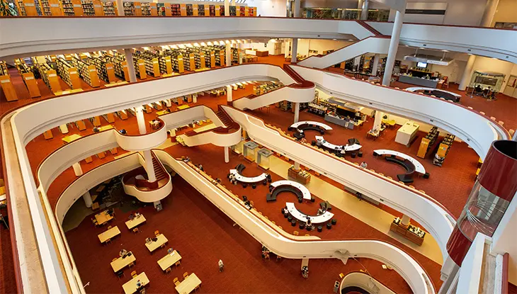Interior de una biblioteca pública con filas de estanterías y zonas de lectura, con una alfombra roja que recorre todo el espacio.