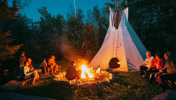 Gens réunis autour d’un feu de camp près d’une tente de style tipi, dans une clairière, éclairés par la chaude lueur du feu au crépuscule.
