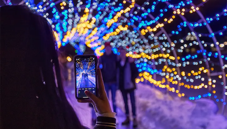 A person takes a photo with their mobile phone of two people posed together under a tunnel of holiday lights, lit up in blue and gold.