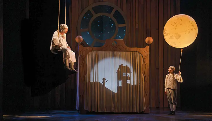 Acteur assis sur une balançoire au-dessus de la scène, tandis qu’un autre tient une lune illuminée.