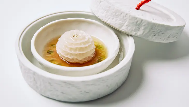 Un plat en forme de fleur dans un bol blanc avec du bouillon, des gouttes d'huile vertes et un couvercle texturé avec une poignée tressée rouge.