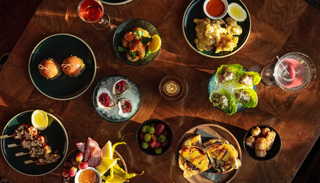 Eine Auswahl an Gourmetgerichten, darunter Austern und Cocktails, auf einem Holztisch im Restaurant Alder im Ace Hotel in Toronto.