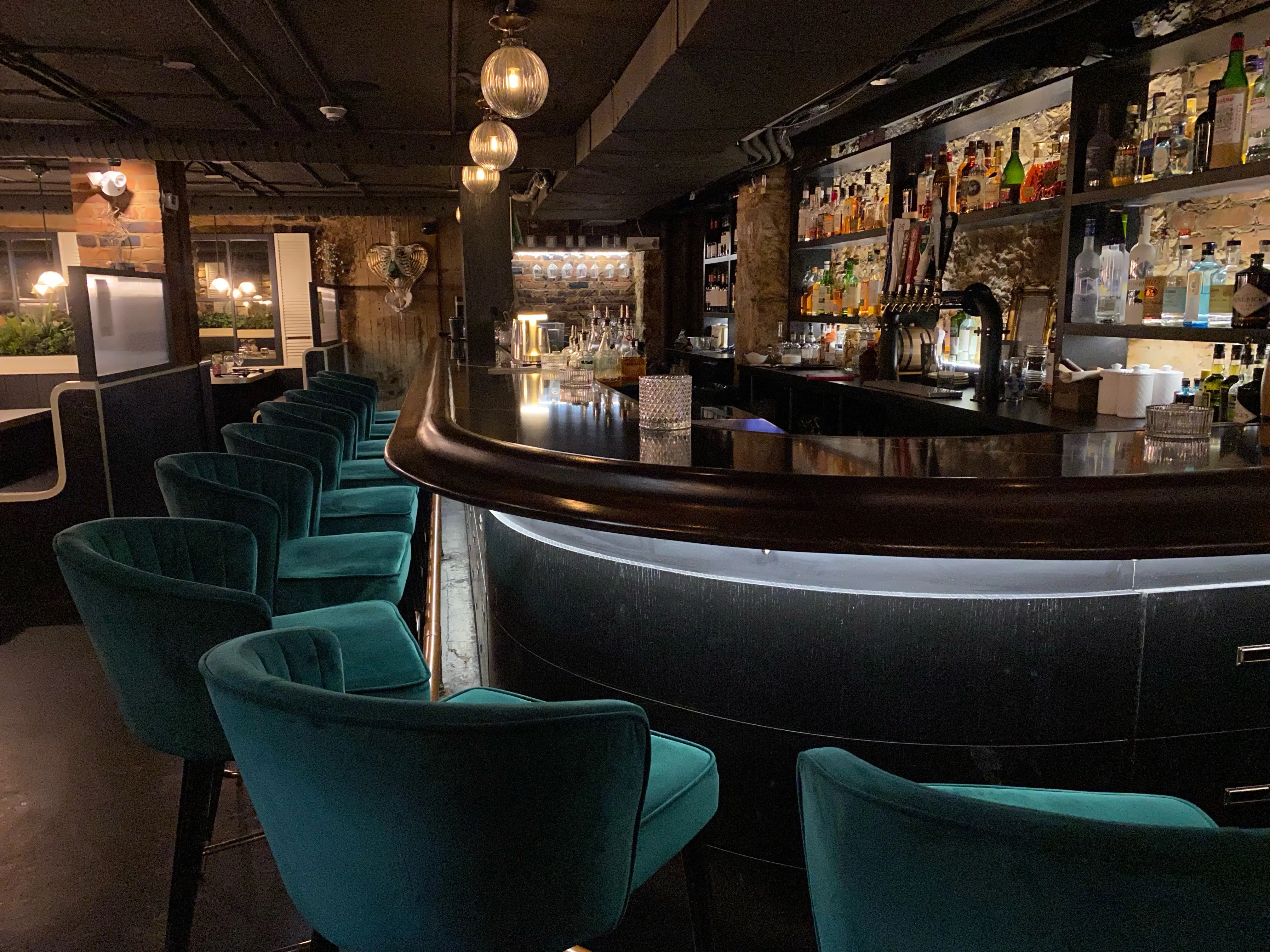 Apothecary Lounge in Ottawa with teal velvet bar stools, a curved dark wood bar and warm ambient lighting in a cozy, upscale setting.