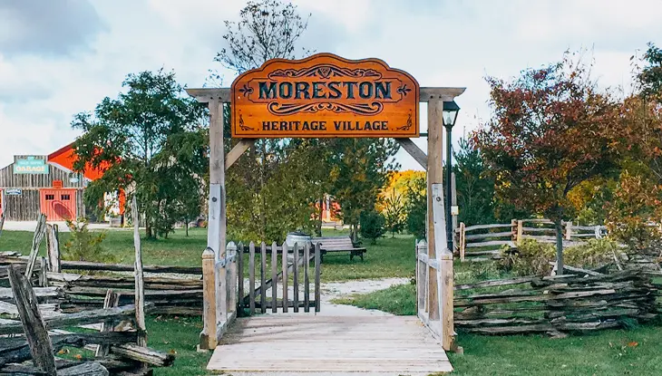 Wooden archway entrance leading into a heritage village with historic fencing and trees.