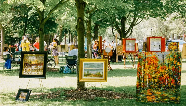 屋外に木々の間に飾られた額入りの芸術作品と、公園を歩く人々の姿。