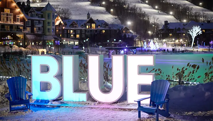 The word "BLUE" in illuminated in large capital letters in blue and white lights at night, at the base of the Blue Mountain ski hill.