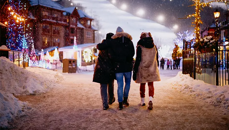 Trois amis se promenant dans le village de Blue Mountain, le soir, avec une pente de ski éclairée devant eux.