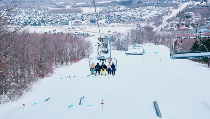 Skiers and snowboarders riding a chairlift over a snow-covered slope, with a winter landscape and residential area visible in the distance.