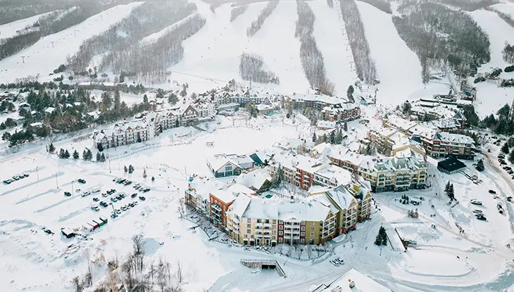 An aerial view of a snow-covered ski resort with slopes, buildings and surrounding forested hills.