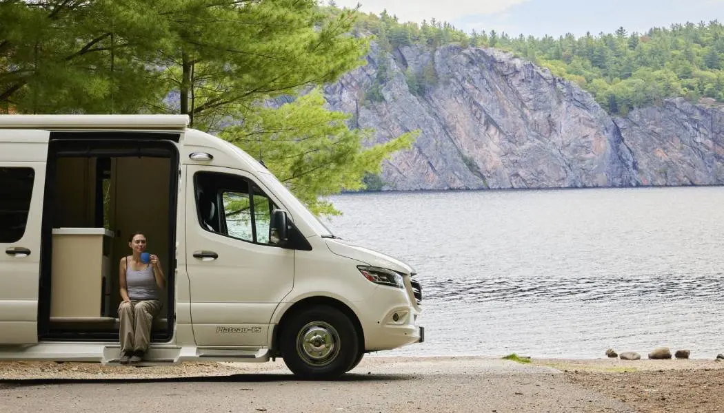 Eine Frau nippt an ihrem Kaffee und sitzt in einem offenen Wohnmobil, das neben einem See im Bon Echo Provincial Park geparkt ist.