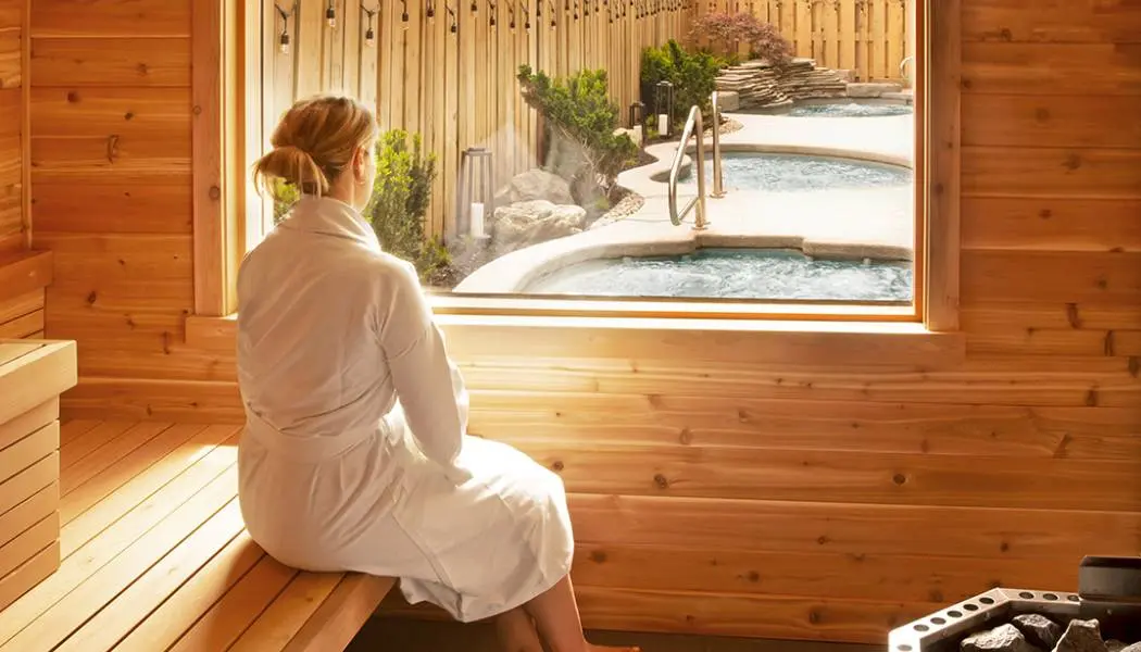 Eine Frau in einem weißen Bademantel sitzt in einer Zedernholzsauna und blickt durch ein Fenster auf die beheizten Außenpools eines Spas.