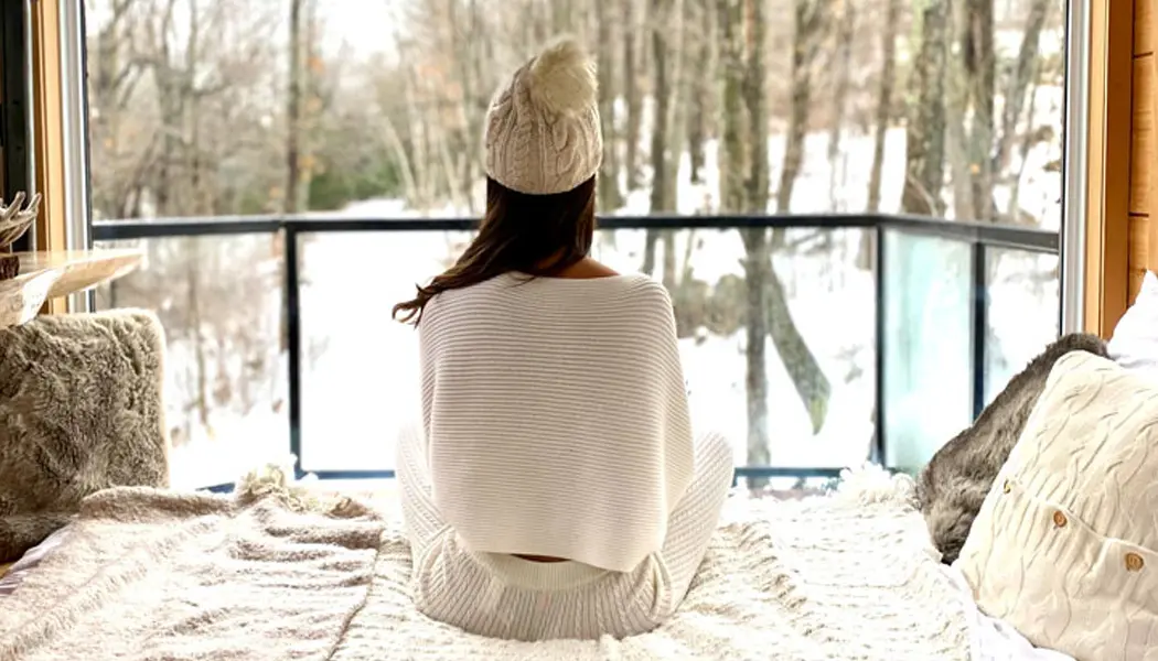 Une femme est assise en tailleur sur un lit confortable, regardant la forêt enneigée à travers une grande fenêtre.
