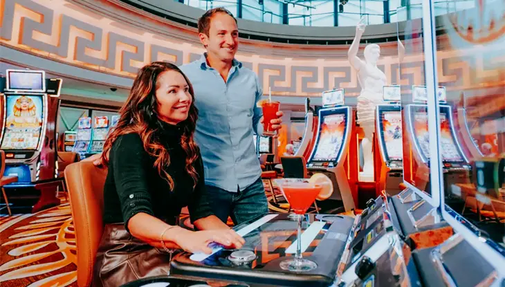 A couple enjoys playing slot machines in a vibrant casino, with colourful decor and drinks.