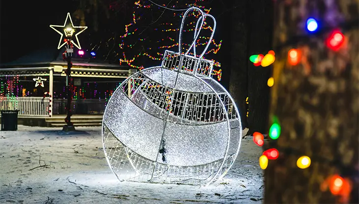 Un gran adorno navideño, creado con luces plateadas, se ilumina por la noche en un parque junto a una glorieta decorada.