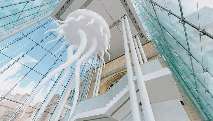 Large white jellyfish-like art installation suspended inside a modern glass atrium with staircases and steel columns.