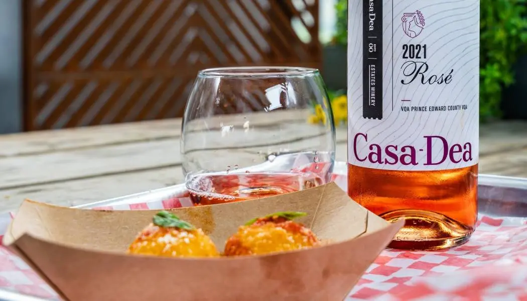 Une bouteille et un verre de vin rosé sont posés sur une terrasse extérieure, à côté d'un plat de canapés à emporter.
