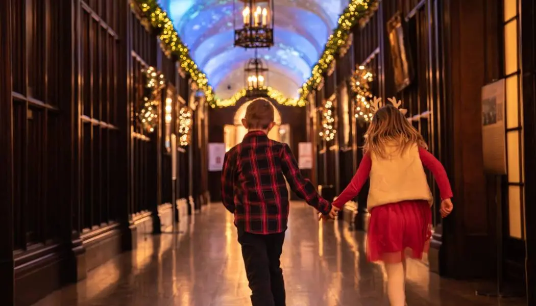 Deux jeunes enfants se tenant par la main et sautillant dans un couloir de Casa Loma, à Toronto, décoré de manière festive pour une activité saisonnière.