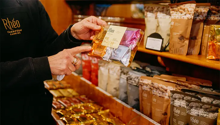 Persona sosteniendo una selección de chocolates envasados ​​dentro de una tienda de dulces, con estantes de dulces variados en el fondo.