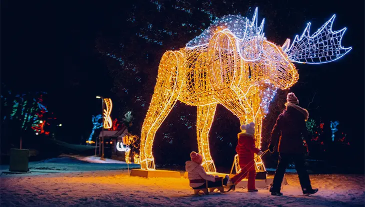 A person pulls a child on a sled, and another child walks beside them, as they pass an illuminated moose structure.