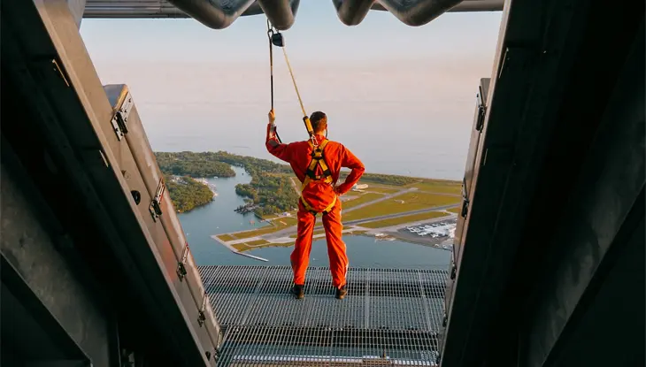 A man wearing a red jumpsuit and safety gear stands on the outdoor platform of EdgeWalk at the CN Tower.