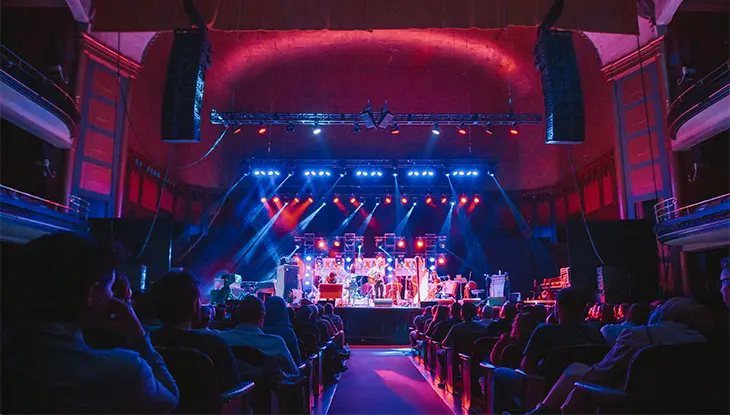 Audience seated inside a darkened concert hall watching a live performance on a brightly lit stage.