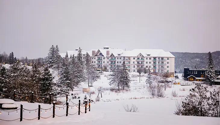 Das Deerhurst Resort in der Abenddämmerung, schneebedeckt, umgeben von Bäumen und sanftem Licht, mit einer friedlichen Winterlandschaft im Blick.