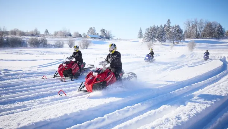 Vier Personen fahren mit Schneemobilen durch eine sonnenbeschienene Schneelandschaft und erzeugen dabei dynamische Spuren im Schnee.