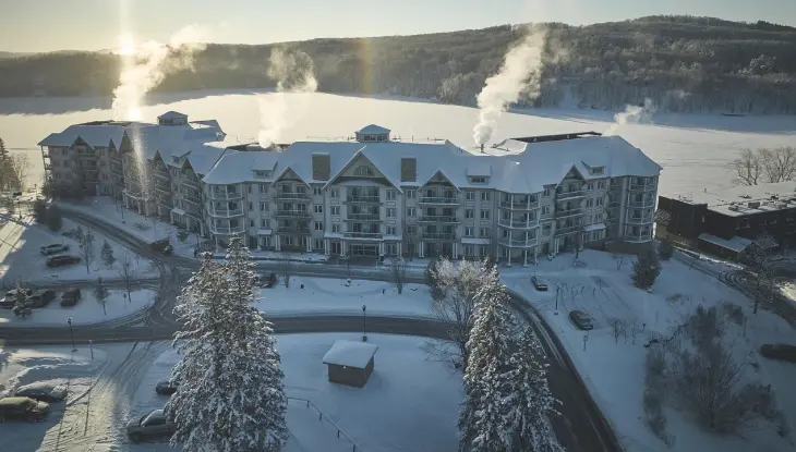 Ein großes Hotel mit schneebedecktem Dach, aus dessen Schornsteinen Rauch aufsteigt, liegt vor der Kulisse eines zugefrorenen Sees und bewaldeter Hügel unter strahlend blauem Himmel.
