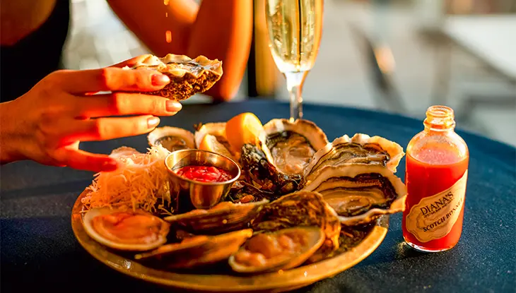 Person holding an oyster over a platter of fresh oysters with lemon, sauce, and a glass of sparkling wine.