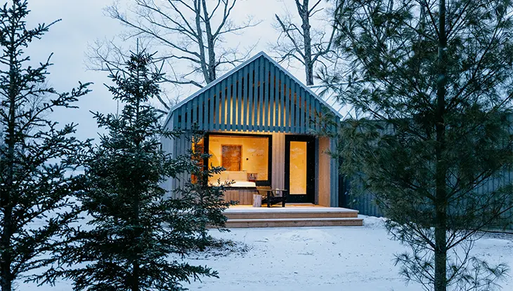 Chalet douillet aux fenêtres illuminées dans une clairière enneigée, entouré de conifères et de feuillus dénudés au crépuscule.