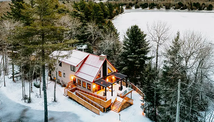 Chalet enneigé et éclairé au milieu d’arbres à feuilles persistantes, près d’un lac gelé.