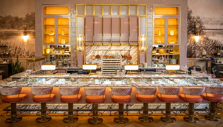 Dining settings are arranged at the marble bar with nine plush stools in a line, facing pink tiled walls behind the bar.