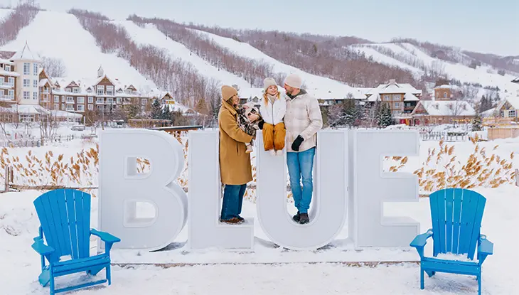 Famille de quatre personnes devant une enseigne du centre Blue Mountain, avec les pentes enneigées et le village en arrière-plan.